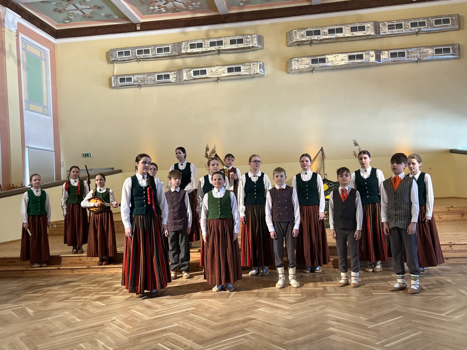 Grupa bērnu un pusaudžu, tērpušies latviešu tautas tērpos, stāv uz koka grīdas zālē. Meitenes valkā baltas blūzes, zaļas vestes un garus svārkus; zēni valkā baltus kreklus, vestes un bikses. Viena meitene tur koklei līdzīgu stīgu instrumentu.