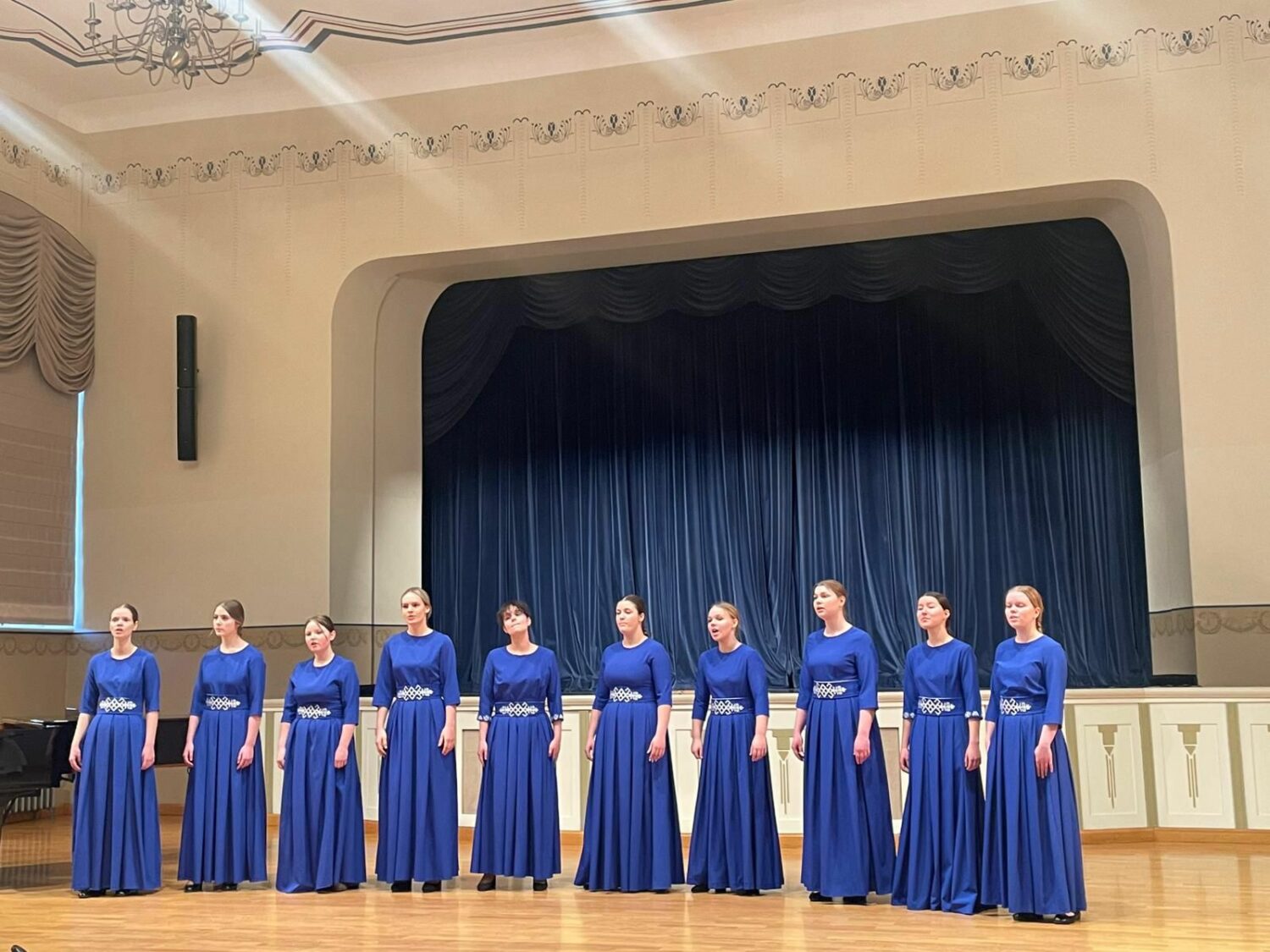 Desmit jaunas sievietes, ģērbušās vienādās garās karaliski zilās kleitās ar sudraba jostām, stāv rindā uz skatuves, raugoties uz priekšu. Aiz viņām ir tumši zili aizkari, un pa kreisi redzama daļa no flīģeļa.