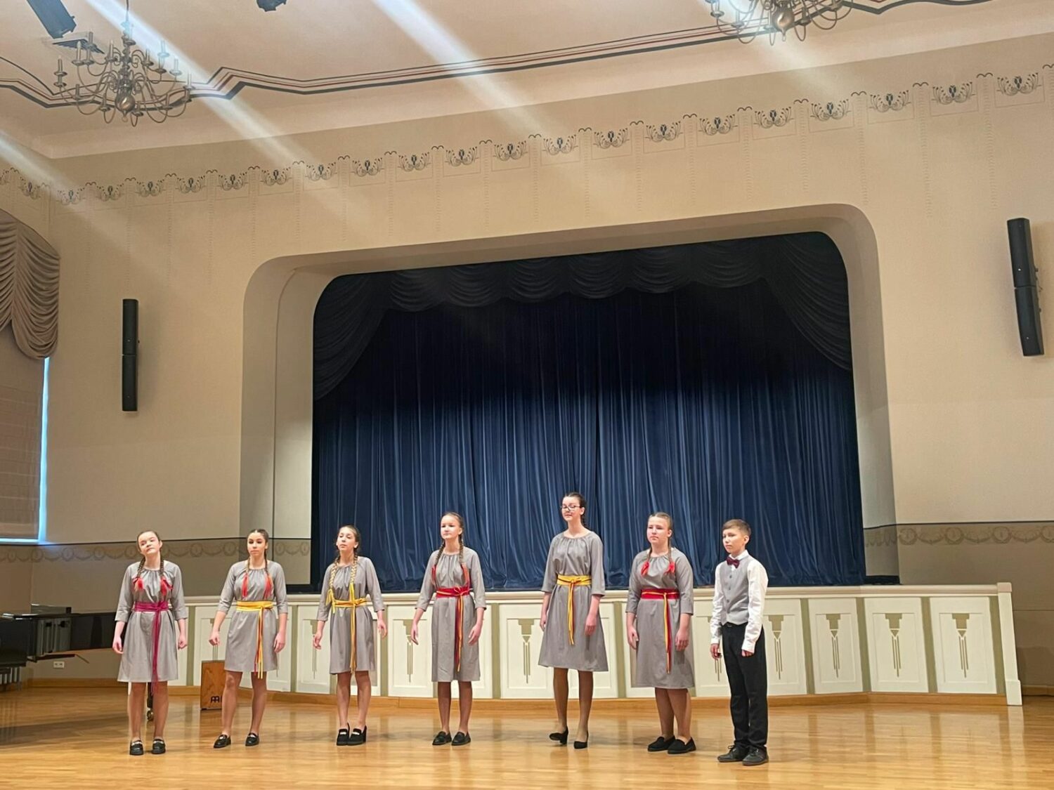 Piecas meitenes pelēkās kleitās ar sarkani dzeltenām jostām un zēns gaišā vestē ar tauriņkaklasaiti stāv uz skatuves ar tumši zilu aizkaru fonā, skatoties uz priekšu.