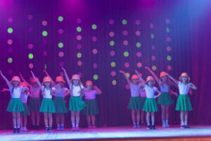 Mūsdienu deju grupa "kENDI" priekšnesums. Mazās dejotājas ar dzeltenām cepurēm un zaļiem svārciņiem.