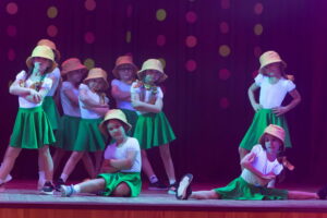 Mūsdienu deju grupa "kENDI" priekšnesums. Mazās dejotājas ar dzeltenām cepurēm un zaļiem svārciņiem.