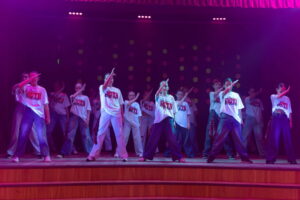 Kendi vecākā grupa dejo uz skatuves, baltos krekliņos ar uzrakstu un džinsās.