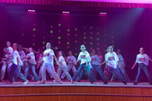 Kendi vecākā grupa dejo uz skatuves, baltos krekliņos ar uzrakstu un džinsās.