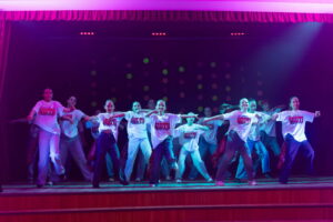 Kendi vecākā grupa dejo uz skatuves, baltos krekliņos ar uzrakstu un džinsās.